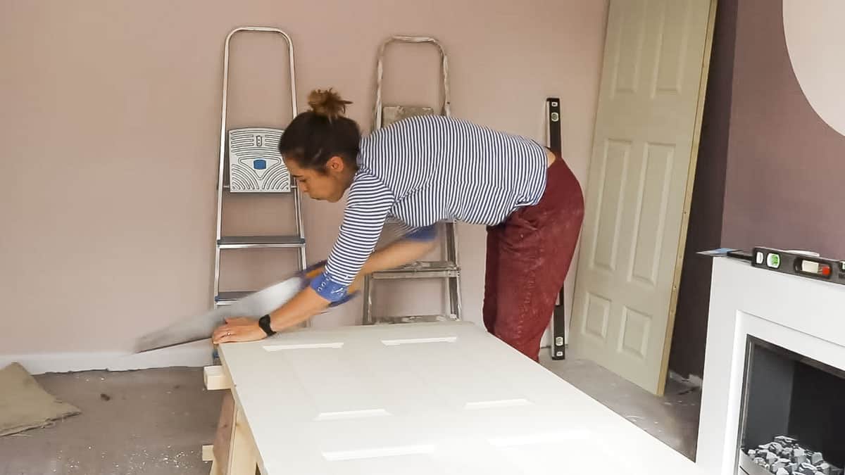 The Carpenter's Daughter cutting an internal door with a hand saw while resting on sawhorses.