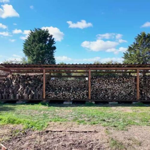 Completed reclaimed timber log store filled with firewood beneath a metal roof beside the existing fence.